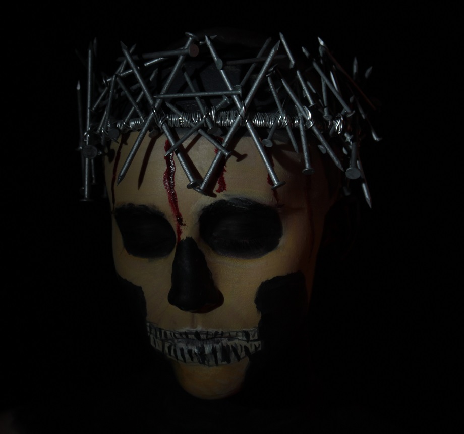 skull face painted while model wears a crown of nails. Stage blood dripping down forehead