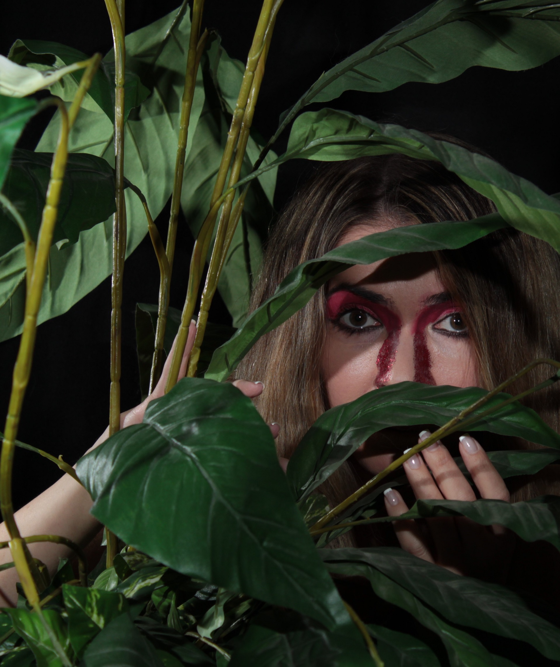 model peaking from behind plant, with red face paint on side of nose and eyelids