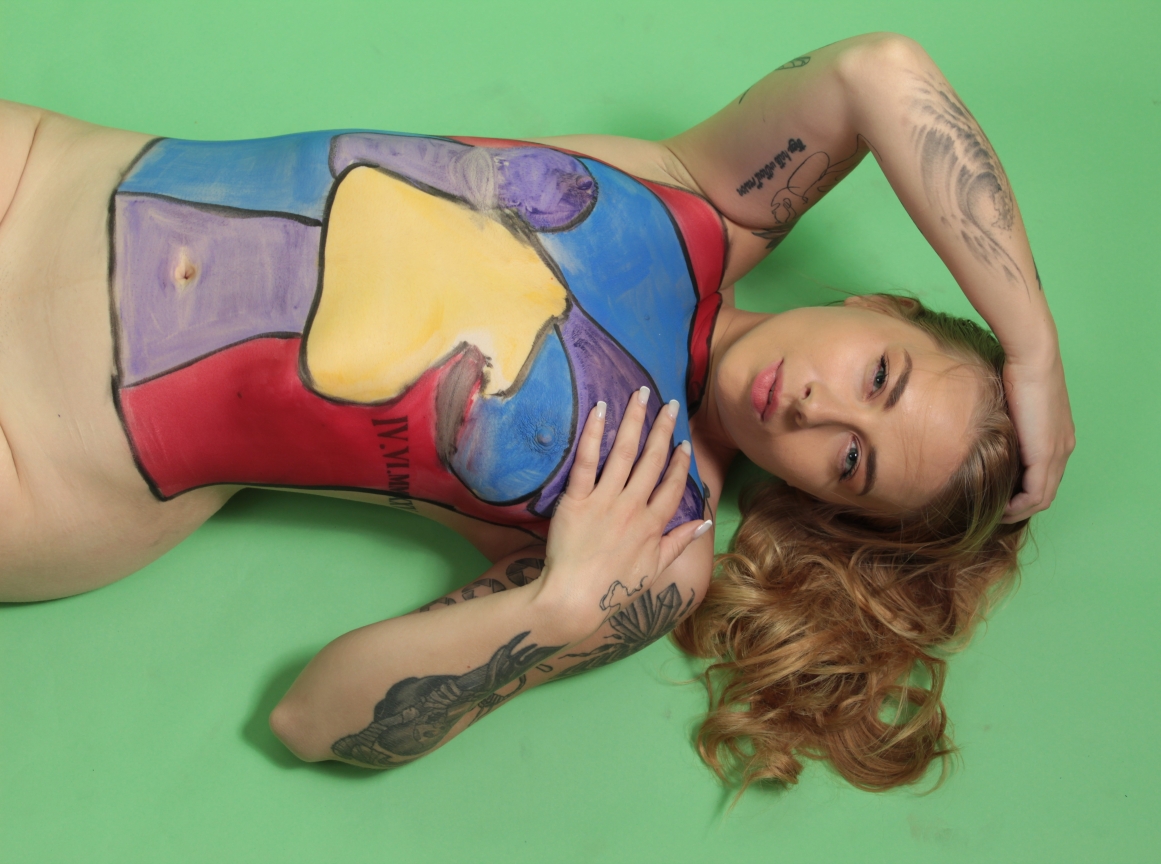 woman laying on green paper backdrop, her torso painted in abstract shapes of blue, red, violet and yellow
