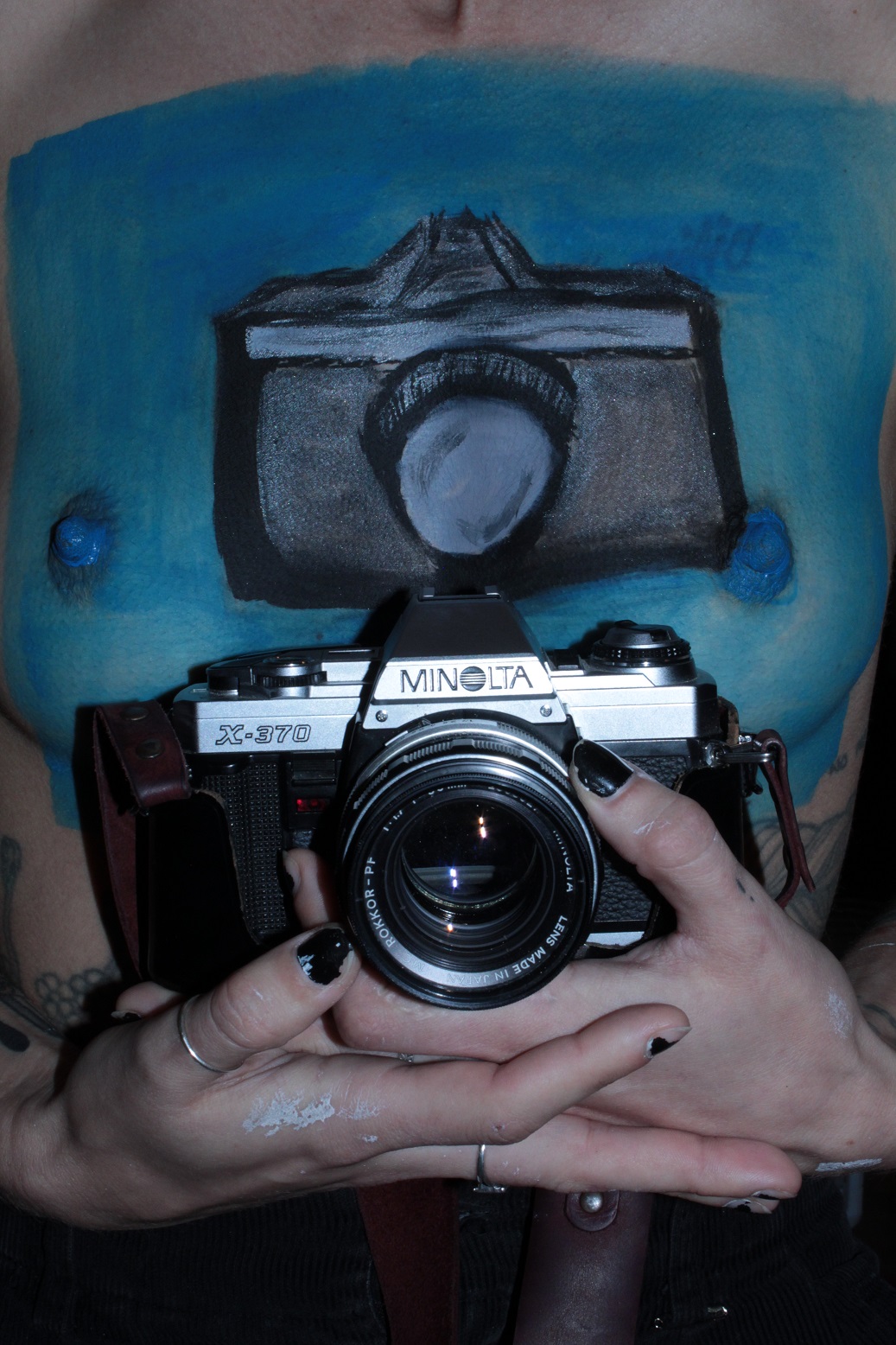 topless female holding a 35-millimeter camera in front her body painted torso with the same camera painted on it